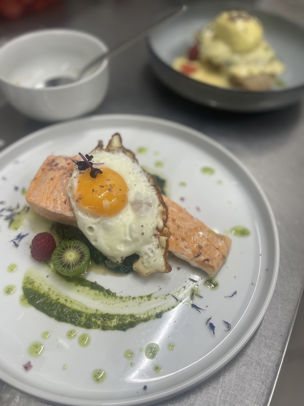 Ein Lachsfilet mit Spiegelei, Mini-Kiwi, Himbeere und grüner Soße auf weißem Teller