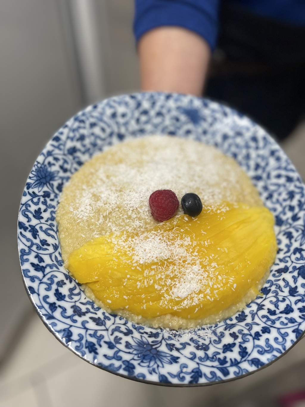 Eine Person hält einen blauen Teller mit hellgelbem Milchreis, Mango, Beeren und Kokosraspeln