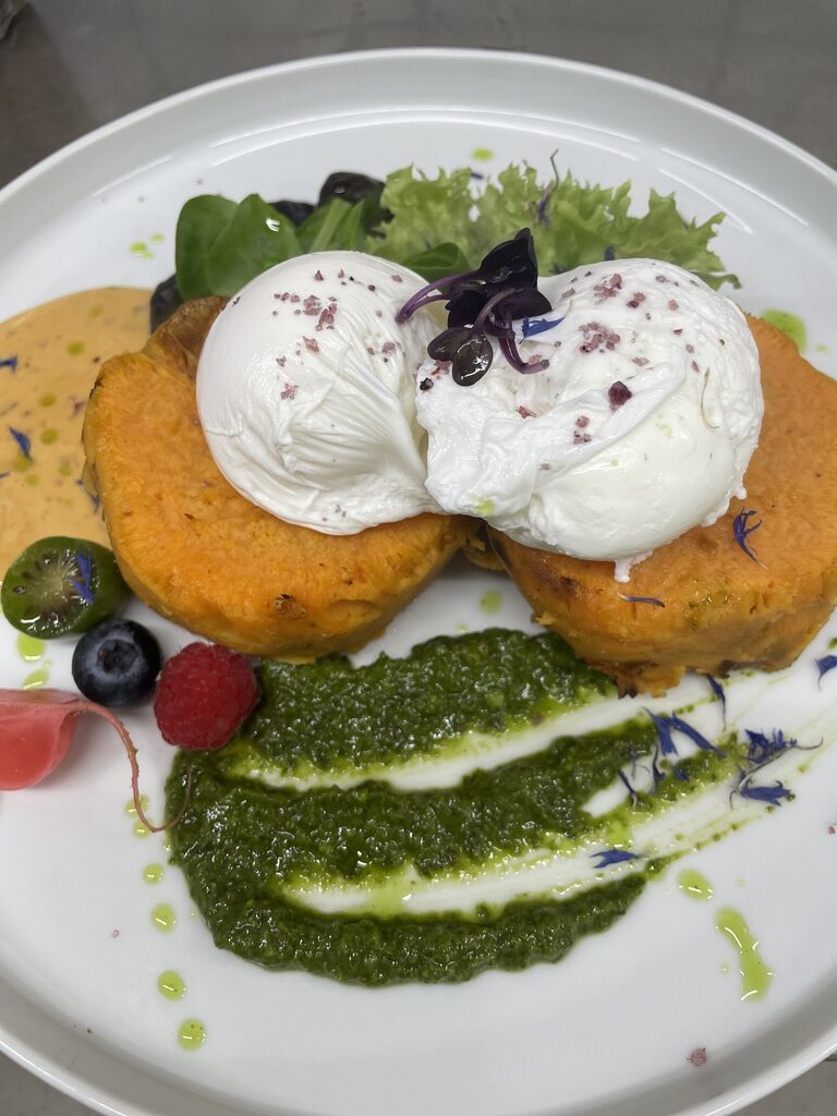 Ein weißer Teller mit zwei pochierten Eiern auf Süßkartoffelscheiben, grünem Pesto, frischen Beeren und farbenfroher Deko