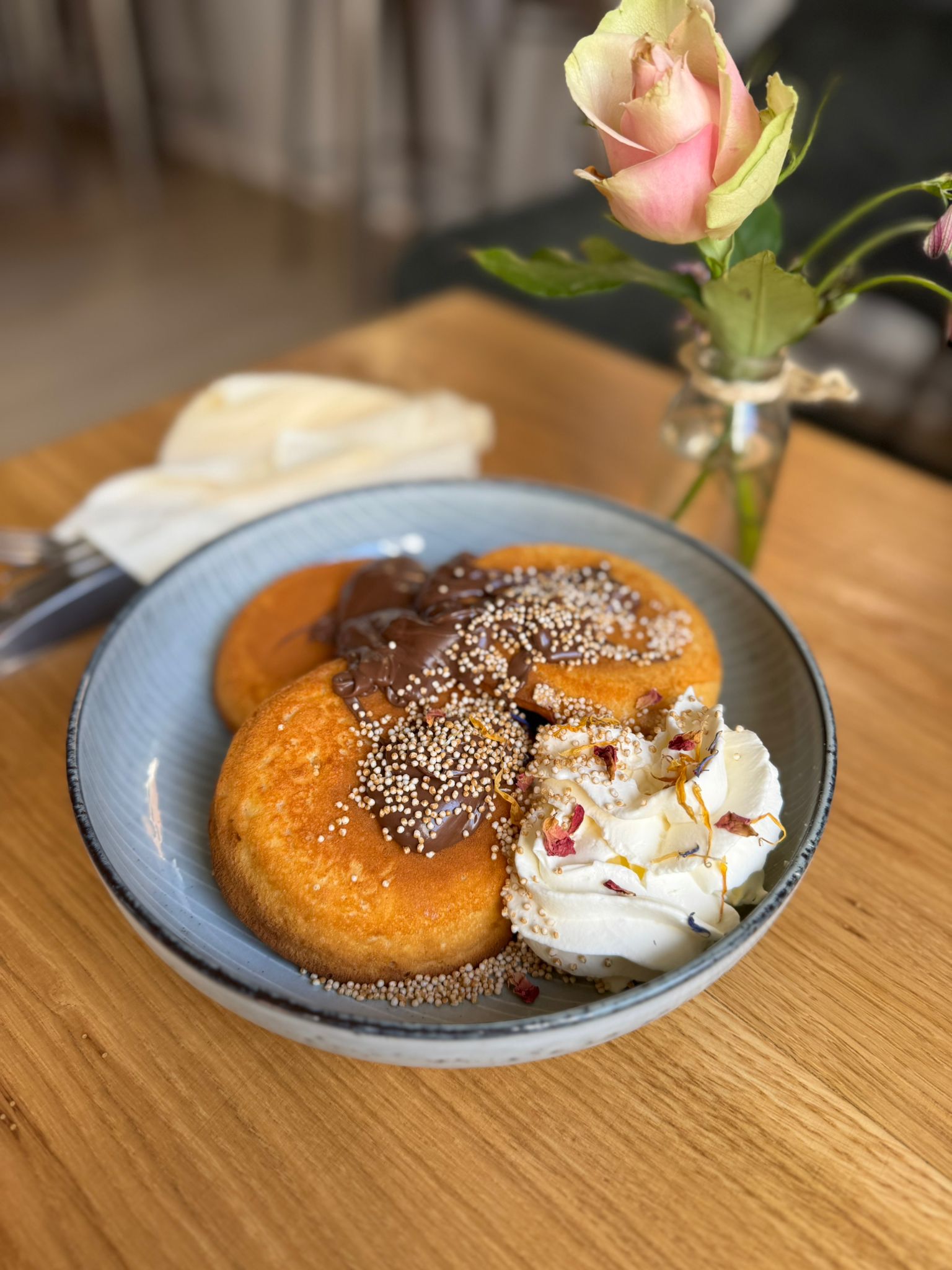Drei goldene Pancakes mit Schokosauce und Sahne auf blauem Teller neben Rose